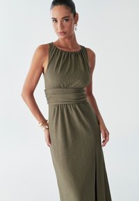 Eine Frau posiert selbstbewusst in einem ärmellosen, olivgrünen Maxikleid mit gerafftem Ausschnitt und einem Seitenschlitz, ergänzt durch Armreifen.