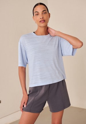 Frau mit dunklem Haar in einem hellblauen Kurzarmshirt und dunkelgrauen Shorts steht mit einer Hand am Nacken in einer neutralen Innenraumumgebung.