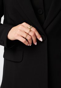 Gouden ringen met een getextureerd draaidesign, een gelaagde ketting en gladde accenten op een hand, die een zwarte blazer draagt.