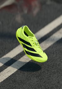 Neon yellow running shoe with black stripes, lightweight design, smooth texture, and lace-up closure, suspended above a road surface.