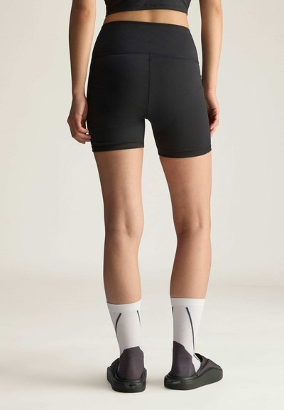 Crne prilagođene kratke hlače s visokim pojasom, glatke teksture i minimalnim šavovima; uparen s bijelim i sivim čarapama sa uzorkom i cipelama za lako obuvanje.