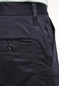 Närbild av marinblå byxor som visar bakfickan med knapp, sid söm, bälteloop och subtila texturen av tyget.