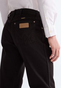 Close-up of a person wearing black Wrangler jeans and a white buttoned shirt, focusing on the back pocket and hand.