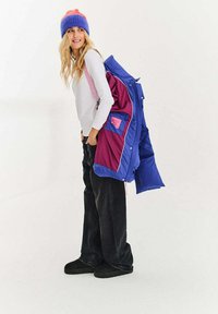 Barevná prošívaná bunda v modré a vínové barvě s fialovými doplňky; nošená přes bílou halenku s dlouhým rukávem a růžovými pásky, černé džíny a boty.