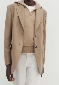 En person iført en beige uld blazer over en tan strikket sweater med krave og off-white bukser foran en ensfarvet lys baggrund.