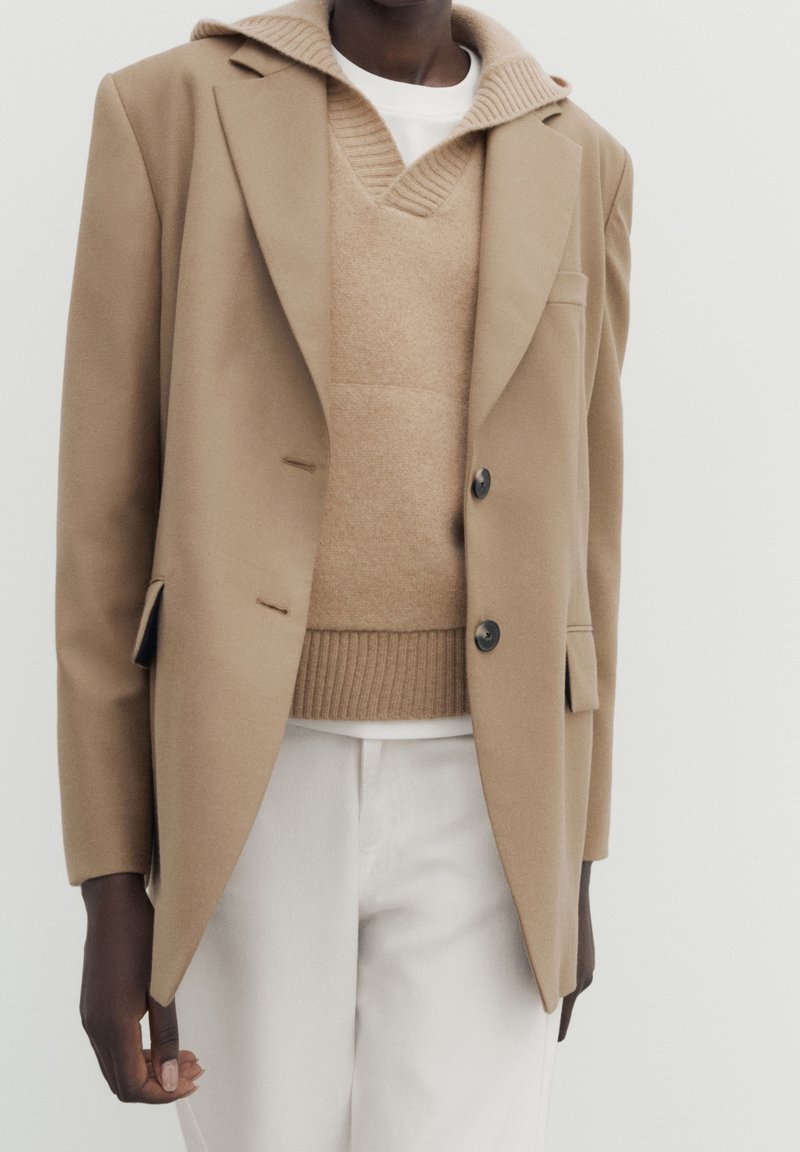 En person iført en beige uld blazer over en tan strikket sweater med krave og off-white bukser foran en ensfarvet lys baggrund.