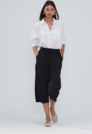 Femme en chemise blanche boutonnée, pantalon large court noir, ballerines blanches tissées et bijoux en or, marchant sur un fond uni.