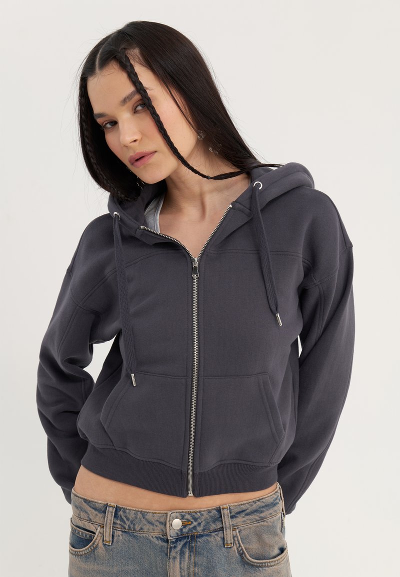 Jeune femme aux longs cheveux foncés, portant un sweat à capuche gris foncé zippé et un jean bleu délavé sur un fond clair uni.