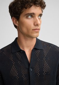 Polo shirt nera in maglia con un motivo a rete e disegni geometrici, due bottoni al colletto e una finitura testurizzata.