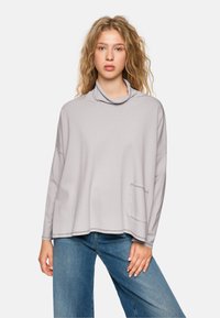 Hellgrauer Pullover mit Kapuze und langen Ärmeln, lockerer Schnitt, ausgestattet mit einer Fronttasche und sichtbaren Nähten. Kombiniert mit blauen Jeans.