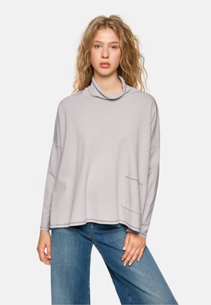 Light grey, cowl neck sweater with long sleeves, relaxed fit, featuring a front pocket and visible stitching details. Paired with blue jeans.