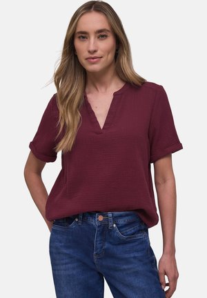 Femme aux longs cheveux blonds portant un chemisier bordeaux à manches courtes et un jean bleu, debout avec une main sur la hanche.