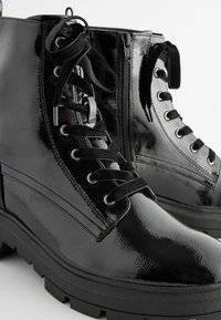 Bottines en cuir verni noir à finition brillante, avec lacets sur le devant, fermeture éclair et semelle texturée antidérapante.