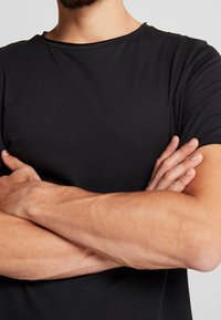 Czarny krótki t-shirt z krótkim rękawem, wykonany z miękkiego materiału, z okrągłym dekoltem. Model ma skrzyżowane ramiona, co ujawnia gładką fakturę.