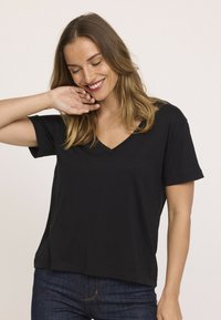 Femme souriante avec des cheveux à longueur d'épaules touche son visage, portant un t-shirt noir à col en V à manches courtes et un jean bleu foncé, sur fond uni.