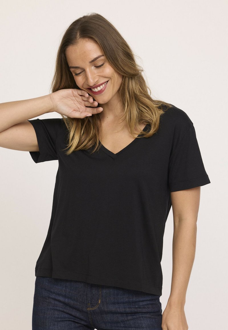 Femme souriante avec des cheveux à longueur d'épaules touche son visage, portant un t-shirt noir à col en V à manches courtes et un jean bleu foncé, sur fond uni.