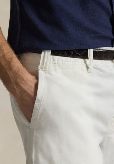 Weiße Baumwollhose mit lockerer Passform, ausgestattet mit Seitentaschen und einem braunen, strukturierten Ledergürtel, der über einem marineblauen Poloshirt an der Taille getragen wird.