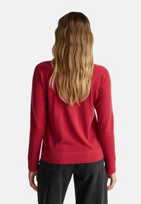 Maglione rosso a maglia con maniche lunghe, polsi e orlo a coste. Presenta una texture liscia. Indossato con pantaloni neri, visto da dietro.