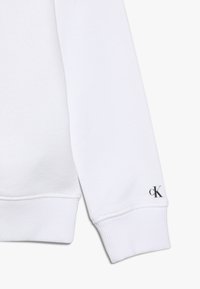 Biała bluza z gładkiego materiału; posiada ściągacze na rękawach i małe czarne logo "cK" na lewym rękawie.