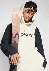 Snowboarder iført en tofarvet (cremefarvet og marineblå) vandtæt jakke med Oakley-logoet, mens han holder en ski med et geometrisk mønster og pink accenter.