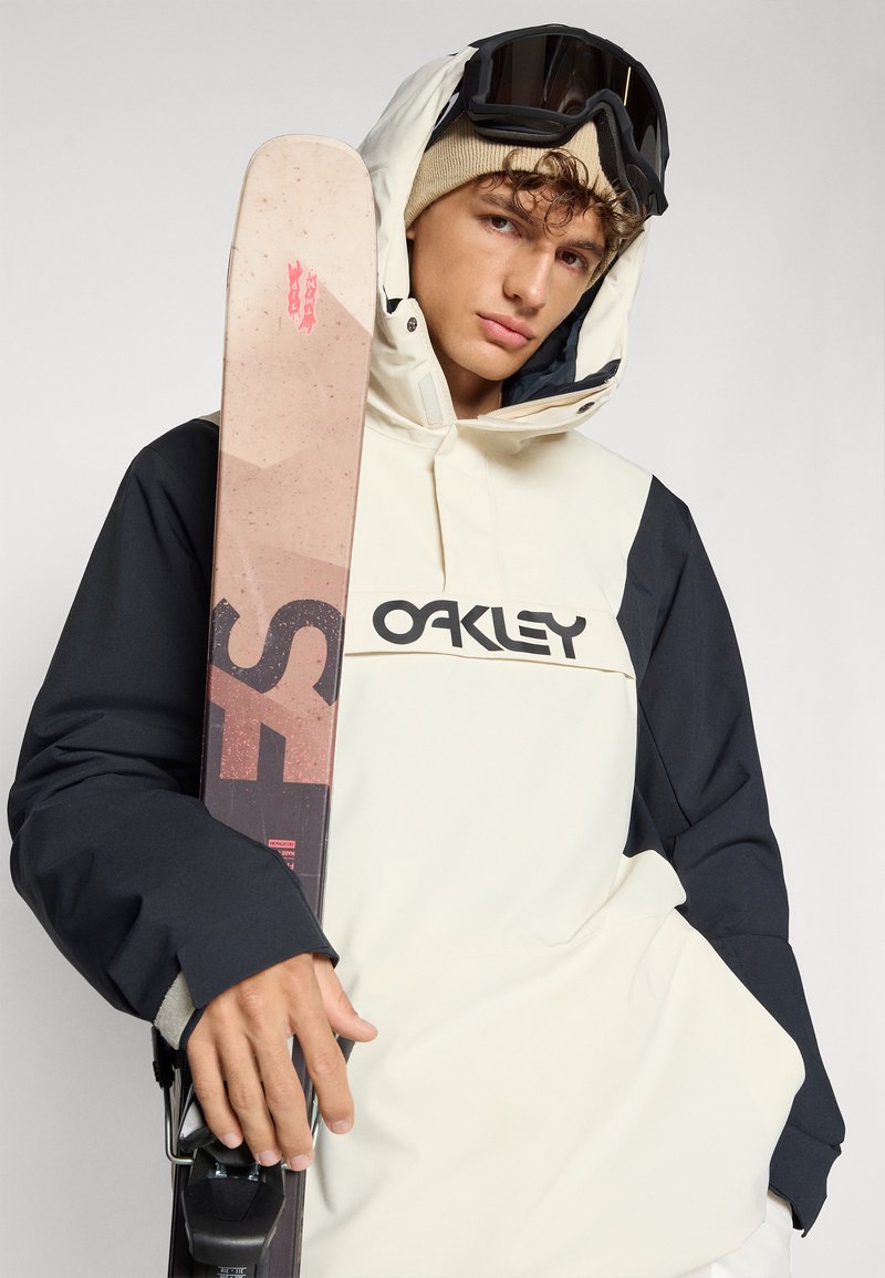 Un snowboarder portant une veste imperméable bicolore (crème et marine) avec le logo Oakley, tenant un ski avec un motif géométrique et des accents roses.