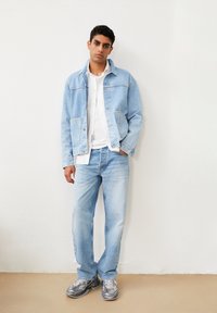 Giubbotto di jeans blu chiaro con tasche a pattina, camicia bianca classica, jeans blu chiaro e sneaker grigie con dettagli testurizzati.