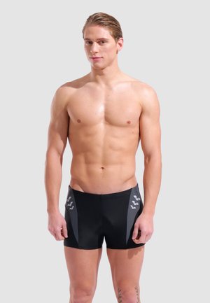 Jeune homme aux cheveux mouillés coiffés en arrière, portant un short de bain noir et gris, debout devant un fond clair et uni.