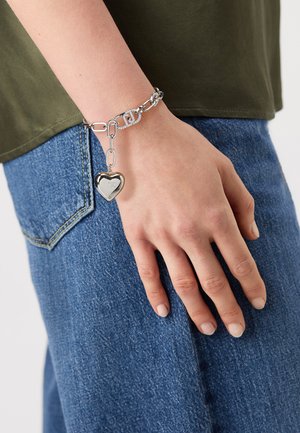 Sølv hjerteanheng armbånd med teksturerte lenker, med et fremtredende hjertevedheng. Båret på en hånd, med en avslappet denim bakgrunn.