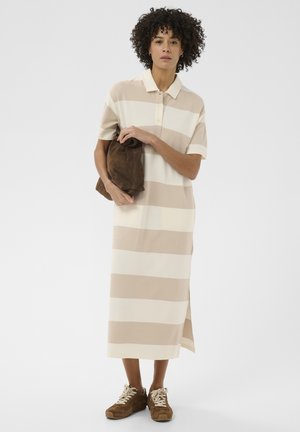 Moteris su garbanotais plaukais dėvi midi ilgio suknią su smėlio ir baltos spalvos juostelėmis, laiko rudą delninę, derina su rudais zomšiniais sportbačiais.