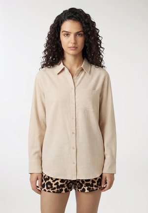 Donna con capelli ricci scuri indossa una camicia beige con bottoni e pantaloncini fantasia leopardata, in piedi contro uno sfondo bianco semplice.