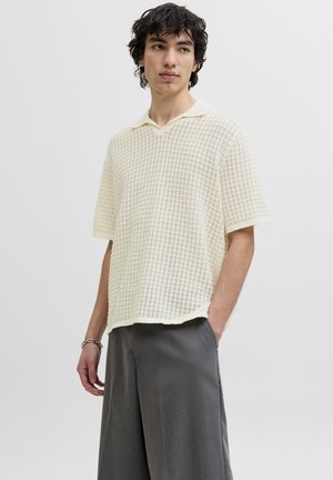 Giovane uomo che indossa una polo a maniche corte color crema con tessuto strutturato e pantaloni grigi larghi, in piedi con una mano in tasca.