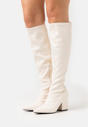 Bottes blanches au-dessus du genou en matériau synthétique lisse et élégant, bouts pointus et talons épais. Dotées d'une fermeture éclair latérale pour faciliter l'enfilage.