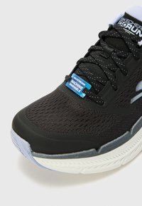 Scarpa sportiva nera con tomaia in rete traspirante, superficie texturizzata, etichetta blu "lavabile in lavatrice" e suola in gomma bianca con motivo per la trazione.
