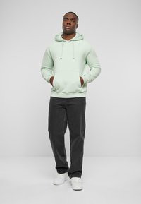 Felpa verde chiaro con tasca a marsupio e cordini regolabili, abbinata a pantaloni grigi scuri e sneakers bianche. Design minimalista.