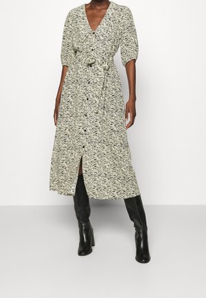 Kvinde iført en lang, beige og sort mønstret kjole med knapper foran, puffede ærmer og bælte i taljen, stylet med sorte knælange støvler med hæl.