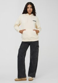 Kremowa bluza z kapturem i kieszenią kangurką, z czarnym logo "CRIME". Połączona z luźnymi czarnymi spodniami cargo i beżowym obuwiem.