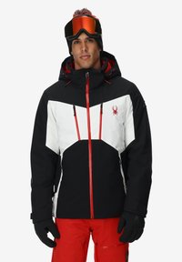 Giacca isolata bianca e nera con accenti rossi, dotata di zip frontale, cappuccio regolabile e multiple tasche con zip. Inclusi guanti e occhiali da sci.