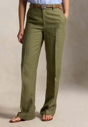 Woman wearing olive green wide-leg trousers with a brown belt and tan sandals, standing against a neutral background.