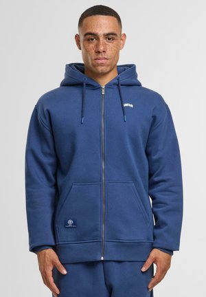 Granatowy hoodie na zamek z przednią kieszenią, kapturem ze sznurkiem, ściągaczami na mankietach i małym logo na piersi. Gładka tekstura bawełnianego materiału.