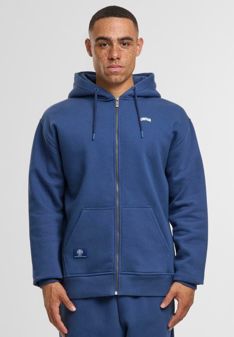 Marineblauwe zip-up hoodie met een voorzak, een capuchon met trekkoorden, ribbelmanchetten en een klein logo op de borst. Glad katoen met een zachte textuur.