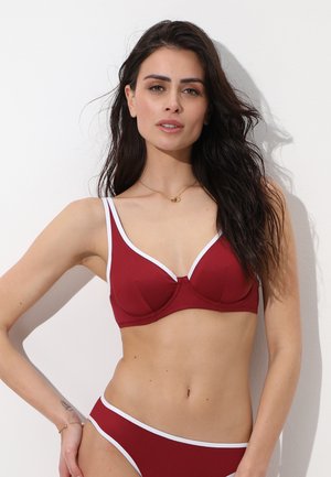 Donna con capelli lunghi e scuri che indossa un bikini rosso con bordi bianchi, in piedi contro uno sfondo bianco con una mano sul fianco.