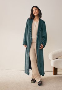 Dlhý tyrkysový zamatový župan s šálovým golierom, v kombinácii s béžovým henley tričkom a zodpovedajúcimi nohavicami. Detaily kvetinového vzoru na čiernych papučiach.