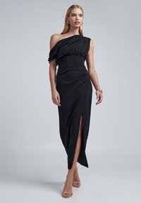 Mustast asümmeetrilisest riidest kleit ühe õlaga, taljesse tõmmatud ja esiosas lõhikuga. Sile tekstuur ja voolav disain. Kaunistatud peente voldikute lisanditega.