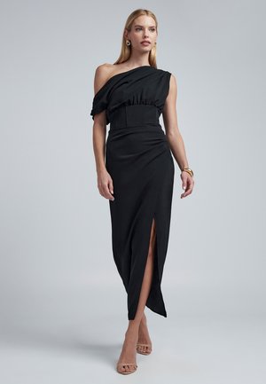 Robe asymétrique noire avec une épaule découverte, taille cintrée et fente sur le devant de la jupe. Texture lisse et design fluide. Accentuée par des drapés subtils.