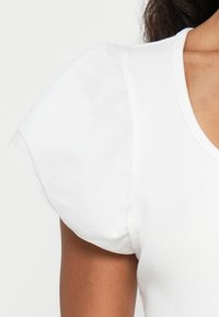 Haut blanc à col en V côtelé avec des manches bouffantes courtes et volumineuses sur le torse supérieur et le bras d'une personne à la peau foncée, devant un fond uni.