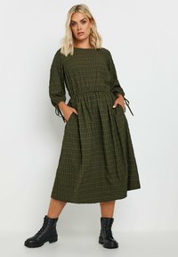 Tummanvihreä ruudullinen mekko, jossa on kolme neljäsosapituista hihaa ja joustava vyötärö. Mekossa on sivutaskut ja rento malli. Yhdistetty mustiin nauhakenkiin.