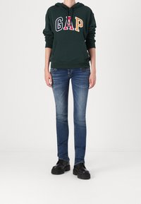 Felpa verde scuro con lettering "GAP" multicolore, abbinata a jeans blu slim-fit e scarpe platform nere. Tasche frontali e cuciture visibili.