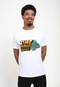 Bílý bavlněný tričko s potiskem kresleného dinosaura s modrými hroty a textem "CESTOVATEL ČASEM Z JINÉHO SVĚTA JURASSIC PORTAL."