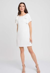 Biała sukienka z krótkim rękawem, z akcentami w postaci kokard na ramionach, o gładkiej fakturze i długości do kolan. Noszona z neutralnymi sandałami na obcasie.