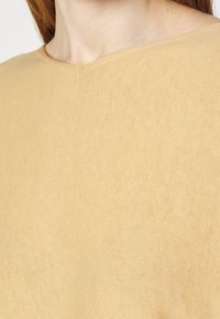 Beżowy top o fakturze z płytkim dekoltem, z prążkowanym wzorem i miękką, lekką tkaniną. Widoczna jest połowa ubrania.